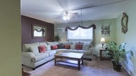 This single-floor space centers around a comfortable and well-appointed living room designed for relaxed everyday living. The room features a large L-shaped sectional sofa in a neutral gray-green upholstery, accented with an assortment of decorative throw pillows in warm red and earth tones. A wooden coffee table with a lower shelf sits at the center, providing both function and visual grounding to the seating arrangement.

A standout design element is the deep brown accent wall, which adds warmth and contrast against the sage green walls throughout the rest of the room. A framed mirror with an ornate gold border is mounted on the accent wall, enhancing the sense of depth and light in the space. Natural light filters in through a broad window dressed with decorative burgundy swag curtains, complemented by a wall-mounted air conditioning unit nearby. An additional window on the adjacent wall further brightens the room.

The ceiling fan with integrated lighting provides both air circulation and ambient illumination. Carpeted flooring adds softness underfoot, while a side table with a classic table lamp offers warm supplementary lighting. The overall ambiance is inviting and lived-in.