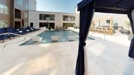 This expansive outdoor amenity space is designed to evoke a resort-like atmosphere, centered around a large rectangular pool that serves as the focal point of the courtyard. The pool features built-in seating ledges with contemporary white lounge chairs positioned directly in the water, alongside integrated water features that add a dynamic element to the setting. The surrounding deck is finished in light travertine tile, lending an upscale, clean aesthetic that complements the modern architectural design of the adjacent building.

The pool deck is well-appointed with multiple seating areas, including cushioned chaise lounges and navy blue recliners arranged along the perimeter. Several private cabanas are distributed throughout the space, each topped with a navy canopy and fitted with flowing curtains that can be drawn for privacy. Inside the cabanas, plush wicker sectional sofas and daybed-style loungers provide comfortable retreats. A set of glass-paneled double doors offers direct interior access from the pool deck, connecting the outdoor space seamlessly to the building's interior. Potted greenery and planters provide subtle landscaping accents. The overall ambiance is polished, social, and ideally suited for relaxed outdoor living.
