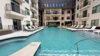 The pool areas depicted showcase two distinct outdoor amenity spaces designed for relaxation and recreation. The first features a expansive, resort-style swimming pool with clear aquamarine water set within a courtyard framed by a contemporary multi-story building with dark brick, large windows, and private balconies. A wide concrete sundeck surrounds the pool, lined with dark-framed chaise lounges topped with white cushions. An adjacent lounge courtyard offers additional seating beneath a modern pergola structure, with manicured plantings and a fire pit adding ambiance.

The second pool setting presents a more traditional aesthetic, characterized by a generously sized rectangular pool accompanied by an adjoining spa. The surrounding deck provides ample space for lounging, with multiple chaise chairs and shade umbrellas scattered throughout. The area is enclosed by multi-story buildings featuring warm wood tones, pitched rooflines, and mature trees that provide natural shade. A covered entryway with rustic wood-framed doors and a posted pool rules sign mark the access point. Sliding glass doors from an adjacent covered patio offer a direct view onto the pool deck, enhancing the sense of indoor-outdoor connectivity throughout the amenity space.