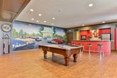 This single-story recreational space in Columbus, Georgia, is designed as a well-appointed gathering and entertainment venue. Warm hardwood flooring flows continuously throughout, unifying the various activity zones within the open layout.

The central focal point of the main lounge area is a dramatic floor-to-ceiling stone fireplace, constructed from natural fieldstone and topped with a mounted flat-screen television. Comfortable seating, including plush sofas and armchairs in rich red and brown tones, is arranged around the hearth to create an inviting atmosphere. Adjacent to this seating area, a regulation-size billiards table anchors the recreational zone, providing ample room for play.

A dedicated kitchenette occupies one end of the space, outfitted with bold red cabinetry, dark countertops, stainless steel appliances including a full-size refrigerator and wall oven, and striking vertical striped wall accents that add a retro-inspired energy.

A quieter seating nook near the entryway features large French doors that bring in natural light and garden views, complemented by upholstered club chairs and framed artwork on the sage-green walls. A separate dining area with a wood table and side chairs offers additional functionality. Recessed lighting and wall sconces provide warm, layered illumination throughout, and decorative murals add personality to the overall design.
