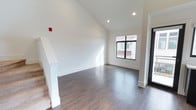 This well-appointed two-story home presents a clean, contemporary aesthetic defined by crisp white walls, vaulted ceilings, and rich dark hardwood-style flooring that flows seamlessly throughout the main level. The open entry area immediately impresses with its soaring ceiling height, which amplifies the sense of space and draws in abundant natural light through a large black-framed window. Recessed lighting complements the natural illumination, ensuring the interior remains bright and welcoming at any time of day. A staircase with a white-painted banister provides a stylish transition between the two floors, serving as both a functional and architectural focal point. The neutral color palette of white walls and warm wood-toned floors creates a versatile canvas that suits a variety of interior styles. The home's modern black-framed entryway door adds a bold, contemporary contrast to the otherwise light interior. The layout is thoughtfully designed to balance open, flowing living areas with defined spaces across both levels, offering a harmonious blend of form and function throughout.