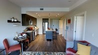This single-floor residence showcases a thoughtfully designed open-concept layout that blends style and functionality. The kitchen is a standout feature, outfitted with dark espresso cabinetry, stainless steel appliances including a gas range, microwave, and refrigerator, and a generous central island with bar seating — ideal for both cooking and casual dining. Rich, dark hardwood flooring extends throughout the main living area, anchoring the space with warmth and cohesion. Recessed lighting and a track light fixture illuminate the kitchen, while the clean white walls and neutral tones create a bright, contemporary atmosphere. The open floor plan allows the kitchen to flow effortlessly into the dining and living areas, fostering a connected and welcoming environment. A hallway visible from the main space leads to additional rooms, hinting at a well-organized and spacious layout beyond the common areas. The overall design aesthetic is modern and polished, with a careful balance of dark and light elements that give the space a refined, urban character. This residence offers a comfortable and visually cohesive living experience throughout its single level.