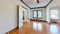 This single-floor space exudes classic architectural character, blending timeless design details with a warm, inviting ambiance. Rich hardwood floors run throughout, anchoring each room with a sense of warmth and continuity. Decorative crown molding and bold dark wood trim frame the ceilings and walls, lending a refined, period-appropriate aesthetic to the interior.

The main living area is generously proportioned, featuring a ceiling fan, a classic cast-iron radiator beneath a double window that frames lush outdoor greenery, and a set of sliding doors that open to adjacent spaces. Natural light flows freely through multiple windows across the unit, brightening each room.

Elegant rounded archways connect the rooms, creating a graceful flow between spaces without sacrificing definition. One room features a textured wall finish and its own radiator beneath a window, while another offers a clean, minimalist feel with a rich dark wood entry door, contrasting trim, and crisp white walls.

A bathroom is also present, identifiable from the overhead view, fitted with classic white fixtures. Throughout the space, the interplay of white walls, dark wood accents, arched openings, and gleaming hardwood floors creates a cohesive, character-rich environment that honors its traditional architectural roots.