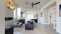 This single-floor space showcases a thoughtfully designed open-concept layout that seamlessly connects the living area to the kitchen. The living space is anchored by light hardwood flooring that runs throughout, creating a warm and cohesive foundation. Crisp white walls and recessed lighting are complemented by a sleek, large-blade ceiling fan, contributing to the modern aesthetic. The living area is furnished with black leather seating arranged around a contemporary low-profile coffee table with gold-tone accents, adding a sophisticated touch. Vertical windows allow natural light to filter in, enhancing the bright and airy atmosphere. The kitchen, visible from the living area, features dark cabinetry and modern appliances, maintaining the space's consistent contemporary palette. Framed artwork adorns the walls, adding personality throughout. A doorway leads to additional areas of the unit. Outside, a generously sized wooden deck features a sleek glass-panel railing system, offering unobstructed views of a tranquil pond and the surrounding landscape — an ideal setting for relaxing or entertaining outdoors.