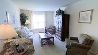 This single-floor space offers a comfortably appointed living area designed with a classic, traditional aesthetic. The living room serves as the centerpiece, featuring plush wall-to-wall carpeting in a soft neutral tone that adds warmth underfoot. Large windows dressed with elegant floral curtains allow generous natural light to fill the room, creating a bright and inviting atmosphere. The space is thoughtfully arranged with a floral-patterned sofa and coordinating armchairs, centered around a wood-and-glass coffee table that provides both style and functionality. A substantial dark wood armoire with decorative ironwork detailing serves as a prominent focal point, offering ample storage and display space, and is topped with lush greenery for a touch of natural elegance. A classic floor lamp provides additional ambient lighting, complementing the room's soft, welcoming feel. The textured ceiling adds subtle architectural character throughout. Overall, the space conveys a relaxed, homey ambiance well-suited for comfortable everyday living and entertaining.