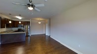 This single-floor space offers an open-concept layout that seamlessly connects the living area to the kitchen, creating a sense of spaciousness and ease of movement throughout. Rich, dark hardwood flooring runs continuously across the main living area, adding warmth and a polished aesthetic to the interior.

The living area is outfitted with a five-blade ceiling fan with an integrated light fixture, providing both comfort and ambient lighting. Neutral-toned walls and clean white baseboards contribute to a bright, modern atmosphere throughout the space.

The kitchen is positioned to one side of the open layout and features dark wood cabinetry with a stainless steel refrigerator and built-in microwave, as well as a dark granite or stone countertop island. The kitchen's design balances functionality with a contemporary style that complements the rest of the space.

A pendant light fixture illuminates a transitional area leading toward an interior door, suggesting access to additional rooms such as a bedroom or bathroom. Recessed vents in the ceiling indicate a central HVAC system for year-round climate control.

Overall, the space presents a clean, move-in-ready environment with consistent finishes, practical fixtures, and an open floor plan well-suited for comfortable everyday living.