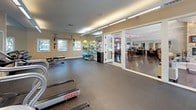 This single-floor fitness center offers a comprehensive workout environment designed to accommodate a wide range of exercise needs. The space is thoughtfully laid out, with distinct zones dedicated to cardio training, strength work, and functional fitness. Rubber flooring throughout provides durability and safety underfoot, while recessed linear lighting along the ceiling ensures consistent, bright illumination across the entire facility.

The cardio zone is anchored by a row of treadmills equipped with individual monitors, alongside elliptical trainers and other aerobic equipment. Full-length mirrors line the walls, visually expanding the space and allowing users to monitor their form during workouts. A dedicated free weights area features a multi-tiered dumbbell rack with a full range of weights and an accompanying flat bench.

The functional training zone is outfitted with stability balls, resistance bands, wall-mounted cable systems, and exercise mats, making it suitable for dynamic and rehabilitative exercise. Natural light enters through multiple windows along the perimeter, enhancing the open, airy atmosphere. Large glass doors connect the fitness area to an adjacent lounge and common space, creating a seamless flow between the two areas and offering views of the surrounding environment.