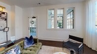 This thoughtfully designed single-floor space seamlessly blends classic and contemporary aesthetics throughout its well-appointed rooms. The living area is anchored by rich, dark hardwood floors and bathed in natural light streaming through a series of large multi-pane windows and a glass-paneled French door that opens to the outdoors. Clean white walls, bold baseboards, and a relaxed, curated furnishing arrangement create an inviting and comfortable atmosphere.

Adjacent to the living space, the dining area features elegant board-and-batten wainscoting, dark hardwood floors, and a striking brass chandelier overhead. A set of French doors with divided glass panes adds architectural interest and connects the room to neighboring spaces.

The kitchen is a standout feature of the home, designed as a generous galley-style layout with bold emerald and teal cabinetry paired with brass hardware and white quartz countertops. A white subway tile backsplash with dark grout lines runs the length of the wall beneath open floating shelves, which display plants and decorative objects. A bridge-style brass faucet over an undermount sink, paired with large windows overlooking lush greenery, adds warmth and character. The iconic black-and-white checkered tile floor carries throughout the kitchen and into the utility and entry areas, lending a retro-modern flair. Globe pendant lights in brushed gold illuminate the space with a warm, welcoming glow throughout.
