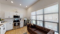 This single-floor unit offers a cohesive blend of modern design and functional living. The open-concept layout seamlessly connects the kitchen and living area, creating an inviting and efficient use of space. The kitchen is outfitted with crisp white cabinetry accented by matte black hardware, white subway tile backsplash, light quartz countertops, and a full suite of stainless steel appliances, including an electric range and over-the-range microwave. Light hardwood-style flooring anchors the kitchen and living area. The living space is bathed in natural light through a large bank of windows fitted with horizontal blinds, offering both brightness and privacy. The bedroom features plush carpeting, clean white walls, and thoughtful built-in window details that allow additional light to filter through. A second bedroom or sleeping area mirrors the same understated, contemporary aesthetic. The bathroom is finished with floor-to-ceiling white subway tile in a generously sized walk-in shower enclosed by a frameless glass door with brushed nickel hardware, lending a clean and polished feel. Recessed lighting throughout the unit ensures a well-lit environment, while the consistent white walls and neutral palette create a bright, airy ambiance across all areas.