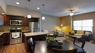 This two-floor residence offers a thoughtfully designed open-concept layout that connects the kitchen, dining, and living areas into a cohesive and functional living space. The kitchen is outfitted with dark espresso cabinetry, stainless steel appliances including a full-size refrigerator, and a granite-topped island that serves as both a workspace and casual gathering spot. Pendant lighting above the island adds a warm, focused glow, complementing the recessed ceiling lights throughout. The dining area accommodates a substantial dark wood table and chairs, making it well-suited for both everyday meals and entertaining. Adjacent to the dining space, the living room features a ceiling fan with integrated lighting, neutral-toned walls with warm yellow accent elements, and ample room for seating. The open sightlines between all three areas create a sense of spaciousness and ease of movement throughout the main floor. Rich hardwood flooring runs throughout the main living area, adding warmth and continuity. The overall aesthetic blends contemporary design with inviting, comfortable details, resulting in a space that is both practical and visually appealing. A second floor provides additional living or sleeping accommodations.