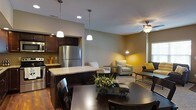 This two-floor residence offers a thoughtfully designed open-concept layout that connects the kitchen, dining, and living areas into a cohesive and functional living space. The kitchen is outfitted with dark espresso cabinetry, stainless steel appliances including a full-size refrigerator, and a granite-topped island that serves as both a workspace and casual gathering spot. Pendant lighting above the island adds a warm, focused glow, complementing the recessed ceiling lights throughout. The dining area accommodates a substantial dark wood table and chairs, making it well-suited for both everyday meals and entertaining. Adjacent to the dining space, the living room features a ceiling fan with integrated lighting, neutral-toned walls with warm yellow accent elements, and ample room for seating. The open sightlines between all three areas create a sense of spaciousness and ease of movement throughout the main floor. Rich hardwood flooring runs throughout the main living area, adding warmth and continuity. The overall aesthetic blends contemporary design with inviting, comfortable details, resulting in a space that is both practical and visually appealing. A second floor provides additional living or sleeping accommodations.