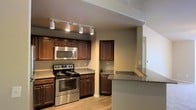 This single-floor space showcases a well-appointed kitchen as its centerpiece, thoughtfully designed to balance practicality with visual appeal. The kitchen features rich, dark wood cabinetry that extends to the upper and lower levels, providing generous storage throughout. Granite countertops with a speckled finish run along the workspace and continue onto a peninsula-style breakfast bar, which offers additional prep space and a natural divider between the kitchen and adjacent areas.

Stainless steel appliances, including a gas range with multiple burners and an over-the-range microwave, lend a contemporary, professional feel to the space. Track lighting mounted on the ceiling provides targeted illumination across the kitchen, enhancing both function and ambiance. The light wood-toned flooring adds warmth and contrasts nicely with the darker cabinetry.

A passthrough or open wall connects the kitchen to the neighboring room, improving the sense of openness and allowing natural light to flow through. Neutral-toned walls complement the overall color palette, creating a cohesive and inviting atmosphere. The layout appears efficient and well-organized, making the most of the available space on this single level.