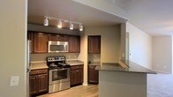 This single-floor space showcases a well-appointed kitchen as its centerpiece, thoughtfully designed to balance practicality with visual appeal. The kitchen features rich, dark wood cabinetry that extends to the upper and lower levels, providing generous storage throughout. Granite countertops with a speckled finish run along the workspace and continue onto a peninsula-style breakfast bar, which offers additional prep space and a natural divider between the kitchen and adjacent areas.

Stainless steel appliances, including a gas range with multiple burners and an over-the-range microwave, lend a contemporary, professional feel to the space. Track lighting mounted on the ceiling provides targeted illumination across the kitchen, enhancing both function and ambiance. The light wood-toned flooring adds warmth and contrasts nicely with the darker cabinetry.

A passthrough or open wall connects the kitchen to the neighboring room, improving the sense of openness and allowing natural light to flow through. Neutral-toned walls complement the overall color palette, creating a cohesive and inviting atmosphere. The layout appears efficient and well-organized, making the most of the available space on this single level.