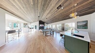 Located at 90 Commons Dr in Eugene, Oregon, this single-floor community lounge is a well-appointed gathering space designed for relaxation and recreation. The room is anchored by a dramatic vaulted ceiling clad in rustic wood planks, lending warmth and architectural character throughout. Recessed lighting and pendant fixtures complement the abundant natural light that pours in through large windows lining multiple walls, offering views of the outdoor pool area.

The open layout centers around a regulation-style pool table positioned prominently on gleaming light-toned hardwood floors. A bold dark-metal fireplace surround rises to the vaulted ceiling peak, framing a mounted flat-screen TV above an electric fireplace, with built-in shelving for décor. A cozy seating area with leather armchairs and a coffee table creates a comfortable lounge zone nearby.

A green kitchen island with bar seating provides a casual refreshment area, while bistro-height tables and bar stools offer additional gathering spots near the windowed wall. University of Oregon spirit is evident in the decorative wall art and signage throughout. Sleek modern accent chairs and an open floor plan complete the space, making it an inviting, energetic environment suited for community socializing.
