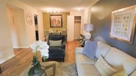 This single-floor residence offers a thoughtfully designed open-concept layout that seamlessly connects the living and dining areas, creating a sense of flow and continuity throughout the space. The living area is anchored by a plush sofa and a coordinating accent chair, complemented by layered area rugs and decorative touches such as an elegant table lamp and fresh floral arrangements that add a refined, lived-in warmth. Warm-toned hardwood flooring runs throughout, unifying the space and enhancing its inviting character. The dining area, set against a rich olive-green accent wall, features a classic chandelier overhead and a well-appointed dining table with upholstered seating, making it an ideal setting for both casual meals and entertaining. A striking framed artwork serves as a focal point on the accent wall, adding personality and depth to the room. A closet with open shelving provides practical storage conveniently situated between the living and dining spaces. The kitchen, partially visible from the main living area, rounds out the functional layout of this well-organized residence. Overall, the space exudes a comfortable and stylish atmosphere, with carefully curated décor and consistent design elements that create a cohesive and welcoming environment.