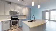 This single-floor space showcases a thoughtfully designed, open-concept layout with a strong emphasis on modern finishes and functional living. The kitchen serves as a central highlight, featuring crisp white quartz countertops that extend across a generously sized island complete with a double stainless steel sink and a contemporary pull-down faucet. Soft blue-grey cabinetry lines the walls from floor to ceiling, providing ample storage while contributing to a calm, cohesive color palette. Stainless steel appliances, including an electric range and over-the-range microwave, add a polished, professional touch. Overhead, a combination of track lighting and elegant cylindrical pendant lights illuminates the space with both task-focused and ambient lighting. The light wood-tone flooring runs throughout, grounding the space and complementing the cool tones of the cabinetry and walls. The open sightlines from the kitchen into the adjoining living area create a seamless flow, making the layout feel connected and spacious. The overall ambiance is clean, bright, and contemporary — ideal for everyday living and entertaining alike.