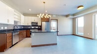 This well-appointed 2-bedroom, 1-bathroom unit offers a thoughtfully designed single-floor layout that maximizes functionality and modern style. The open-concept kitchen serves as the heart of the home, showcasing a two-tone cabinetry scheme with warm wood lower cabinets and crisp white upper cabinets, all accented with gold hardware. A textured tile backsplash, dark stone countertops, stainless steel appliances — including a gas range, microwave, and refrigerator — and a central island with additional storage complete the kitchen. A striking multi-globe brass pendant chandelier adds a contemporary focal point overhead. Light marble-style tile flooring flows throughout the main living areas. The living room features large sliding glass doors and floor-to-ceiling windows that fill the space with natural light and provide outdoor views, complemented by a built-in fireplace with a wood mantel. The two bedrooms are carpeted for warmth and comfort, each with windows offering outdoor views, and one includes a dedicated closet with solid wood bifold doors. The full bathroom features a soaking tub and shower combination with floor-to-ceiling grey tile surround and matte black fixtures. An in-unit stacked washer and dryer add everyday convenience.