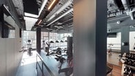 This contemporary fitness center is designed with both functionality and aesthetics in mind. The space features an industrial-style ceiling with exposed ductwork, ventilation pipes, and linear pendant lighting that adds a bold, urban character to the environment. Dark, neutral tones throughout the walls and flooring create a focused, high-energy atmosphere well-suited for exercise.

The gym floor is outfitted with a comprehensive range of equipment, including treadmills, elliptical machines, stationary bikes, and a multi-station cable and strength training system, offering users a full spectrum of cardio and resistance training options. The rubber-matted flooring provides both safety and durability underfoot.

Large floor-to-ceiling windows line one side of the space, offering views of the surrounding streetscape and allowing abundant natural light to pour in — creating an open, energizing environment despite the space's dark interior palette. The expansive glazing also gives the fitness area a sense of openness and connectivity to the outdoors.

Overall, the space strikes a balance between an industrial design aesthetic and a modern, well-appointed gym facility, making it a practical and visually compelling environment for fitness activities.