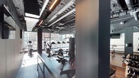 This contemporary fitness center is designed with both functionality and aesthetics in mind. The space features an industrial-style ceiling with exposed ductwork, ventilation pipes, and linear pendant lighting that adds a bold, urban character to the environment. Dark, neutral tones throughout the walls and flooring create a focused, high-energy atmosphere well-suited for exercise.

The gym floor is outfitted with a comprehensive range of equipment, including treadmills, elliptical machines, stationary bikes, and a multi-station cable and strength training system, offering users a full spectrum of cardio and resistance training options. The rubber-matted flooring provides both safety and durability underfoot.

Large floor-to-ceiling windows line one side of the space, offering views of the surrounding streetscape and allowing abundant natural light to pour in — creating an open, energizing environment despite the space's dark interior palette. The expansive glazing also gives the fitness area a sense of openness and connectivity to the outdoors.

Overall, the space strikes a balance between an industrial design aesthetic and a modern, well-appointed gym facility, making it a practical and visually compelling environment for fitness activities.
