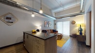 Located at 320 W Broadway in Monona, Wisconsin, this single-floor unit showcases a compelling industrial-modern aesthetic throughout. The open-concept kitchen and living area serves as the heart of the space, where soaring ceilings with exposed silver ductwork and track lighting create a loft-like atmosphere. The kitchen is outfitted with light wood cabinetry, dark granite countertops, black appliances, and a central island with bar seating — ideal for both cooking and casual dining. Pendant lights hang overhead, adding warmth to the modern finishes. Hardwood-style flooring flows seamlessly into the living area, which is anchored by a large sliding glass door that draws in natural light. The bedroom offers a comfortable and private retreat with a window, wall-mounted TV, and a sizable walk-in closet equipped with wire shelving and hanging rods for ample storage. The bathroom continues the granite and light wood cabinetry theme, featuring a single vanity with an undermount sink and a large mirror. A dedicated laundry area houses a stacked washer and dryer unit, adding everyday convenience. The overall design balances raw industrial elements with contemporary finishes for a polished, functional living environment.