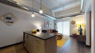 Located at 320 W Broadway in Monona, Wisconsin, this single-floor unit showcases a compelling industrial-modern aesthetic throughout. The open-concept kitchen and living area serves as the heart of the space, where soaring ceilings with exposed silver ductwork and track lighting create a loft-like atmosphere. The kitchen is outfitted with light wood cabinetry, dark granite countertops, black appliances, and a central island with bar seating — ideal for both cooking and casual dining. Pendant lights hang overhead, adding warmth to the modern finishes. Hardwood-style flooring flows seamlessly into the living area, which is anchored by a large sliding glass door that draws in natural light. The bedroom offers a comfortable and private retreat with a window, wall-mounted TV, and a sizable walk-in closet equipped with wire shelving and hanging rods for ample storage. The bathroom continues the granite and light wood cabinetry theme, featuring a single vanity with an undermount sink and a large mirror. A dedicated laundry area houses a stacked washer and dryer unit, adding everyday convenience. The overall design balances raw industrial elements with contemporary finishes for a polished, functional living environment.