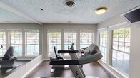 This single-floor community facility is thoughtfully designed to support both recreation and relaxation. The fitness center is well-equipped with Matrix treadmills and a stationary bike, set against dark flooring and textured ceilings. Large windows along one wall provide ample natural light and offer views of the outdoor pool area, while wall-mounted mirrors visually expand the space. A dedicated changing room is accessible directly from the gym floor.

The main lounge area features vaulted, wood-paneled ceilings with a ceiling fan, creating an airy, open atmosphere. A wall-mounted television, fireplace, and designated computer workstations make this a versatile gathering and activity space. Tile flooring transitions seamlessly into carpeting, and wide windows fitted with horizontal blinds allow for controlled natural light throughout.

A screened porch connects the interior to the outdoors, furnished with lounge chairs and a small side table — ideal for casual seating. This leads to a generously sized covered outdoor patio with blue carpeting and open sides, providing shaded views of the fenced pool area, complete with lounge chairs, umbrellas, and mature trees in the background. Together, these spaces create a comprehensive and inviting community amenity center.