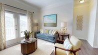 This thoughtfully designed single-floor residence offers a refined living environment that balances classic architectural elegance with contemporary interior styling. The living area serves as the heart of the home, showcasing crown molding that frames the ceiling and crisp white walls that create a bright, airy backdrop. Natural light filters generously through the windows, dressed with patterned drapery in warm gold and blue tones that add a touch of sophistication. The space is furnished with a plush blue velvet sofa accented by an assortment of decorative pillows in gold and neutral tones, complemented by a striped accent chair that reinforces the room's cohesive color palette. A rich wood side table and a round wooden coffee table ground the seating arrangement, while gold-toned sculptural lamps provide warm ambient lighting. A large landscape painting in soft sky blues and greens anchors the main wall, adding a serene focal point. Lush greenery introduces a natural element that enlivens the overall composition. The soft carpeted flooring adds warmth and comfort underfoot, tying together the room's welcoming and polished aesthetic throughout.