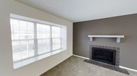This two-floor space offers a thoughtfully designed interior with a welcoming living area as a central highlight. The living room features a striking dark taupe accent wall that draws attention to a classic fireplace, complete with a dark tile surround, matching tile hearth, and a clean white mantel shelf that provides a simple yet elegant focal point. The carpeted flooring adds warmth and comfort underfoot, complementing the neutral tones throughout the space.

Natural light plays a significant role in the living area, provided by a large, multi-pane picture window fitted with horizontal blinds, offering both brightness and privacy control. The surrounding walls are finished in a crisp off-white, creating a balanced contrast against the darker accent wall. The ceiling is smooth and white, contributing to an open, airy feel.

Spanning two floors, the property provides versatile living across multiple levels, offering separation between common areas and private spaces. The overall aesthetic is neutral and understated, providing a clean canvas adaptable to a variety of interior styles and furnishing choices.