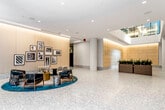 This thoughtfully designed lobby presents a refined, contemporary aesthetic anchored by polished terrazzo flooring that extends throughout the entire space. The reception area commands attention with a sculptural marble-and-black desk set against warmly lit, wood-paneled walls finished in a soft, honey-toned palette. Recessed cove lighting runs along the ceiling perimeter, casting an inviting ambient glow that complements natural light filtering in through large floor-to-ceiling glazed entry doors and an overhead skylight atrium.

A dedicated lounge area offers a curated arrangement of upholstered armchairs and rounded accent tables atop a teal area rug, with a gallery-style collection of black-and-white architectural photography adorning the paneled walls nearby. Potted greenery and planter boxes add organic warmth to the open, column-punctuated floor plan.

The elevator bank features sleek black-framed doors paired with full-height mirrors and matching terrazzo flooring, maintaining a consistent design language throughout. The mailroom is distinguished by bronze individual mailboxes mounted along gold-paneled cabinetry beneath a decorative hand-painted mural, adding an artistic focal point. Together, these elements create a polished, welcoming arrival experience.
