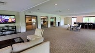 This single-floor facility offers a thoughtfully designed communal space that blends comfort and functionality across several interconnected areas. The layout is anchored by a generous main hall with plush carpeting, recessed lighting, and large floor-to-ceiling windows that invite abundant natural light and views of the surrounding greenery. The open-plan design flows seamlessly between distinct zones, including a welcoming lounge area furnished with wicker-style armchairs arranged around a glass-topped coffee table atop a decorative area rug—ideal for relaxed conversation and social gatherings. Large glass-panel partitions and wide doorways connect the main hall to adjacent rooms, including a dining area outfitted with dark dining tables and chairs, and a secondary lounge featuring additional seating and garden views. Lush indoor plants are placed throughout, adding warmth and a natural ambiance to the interiors. The overall aesthetic is polished yet approachable, with neutral tones, light-colored walls, and tasteful décor that create a calm, inviting atmosphere. Recessed ceiling lights provide consistent illumination throughout, complementing the natural light that fills the space during the day.