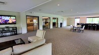 This single-floor facility offers a thoughtfully designed communal space that blends comfort and functionality across several interconnected areas. The layout is anchored by a generous main hall with plush carpeting, recessed lighting, and large floor-to-ceiling windows that invite abundant natural light and views of the surrounding greenery. The open-plan design flows seamlessly between distinct zones, including a welcoming lounge area furnished with wicker-style armchairs arranged around a glass-topped coffee table atop a decorative area rug—ideal for relaxed conversation and social gatherings. Large glass-panel partitions and wide doorways connect the main hall to adjacent rooms, including a dining area outfitted with dark dining tables and chairs, and a secondary lounge featuring additional seating and garden views. Lush indoor plants are placed throughout, adding warmth and a natural ambiance to the interiors. The overall aesthetic is polished yet approachable, with neutral tones, light-colored walls, and tasteful décor that create a calm, inviting atmosphere. Recessed ceiling lights provide consistent illumination throughout, complementing the natural light that fills the space during the day.