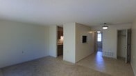 This single-floor residence offers a straightforward and functional layout designed for comfortable everyday living. The main living area is spacious and open, featuring neutral-toned carpet flooring and light beige walls that create a clean, understated backdrop. A textured ceiling adds a classic touch throughout the space. The living area flows naturally toward a tiled entryway and hallway, which is brightened by a window that allows natural light to filter in, enhancing the overall sense of openness.

Partially visible from the main living area, the kitchen features warm, dark wood cabinetry and under-cabinet lighting, offering a cozy contrast to the lighter tones of the surrounding rooms. A pass-through or recessed opening in the wall connects the kitchen area to the main living space, promoting a sense of connectivity between the two areas.

A pendant light fixture is visible near the dining or entry area, adding a simple decorative element. The hallway beyond the main space leads to additional rooms, suggesting a well-organized floor plan. Overall, the space presents a practical and livable environment with a neutral palette and classic residential finishes throughout.