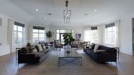 Located at 1600 Club Drive in Woodbury, New Jersey, this single-floor space offers a welcoming and well-appointed communal environment designed for comfort and socializing. The main lounge area is generously proportioned, featuring an open layout that fosters a sense of spaciousness and ease of movement throughout. A standout geometric pendant light fixture anchors the center of the room, drawing the eye upward toward a smooth, white ceiling accented by recessed lighting that provides warm, even illumination throughout the space. Large windows fitted with neutral Roman shades line the walls, inviting an abundance of natural light while offering views of the surrounding greenery. The seating arrangement is thoughtfully composed, with plush gray sofas flanking a sleek, metal-framed coffee table at the heart of the room. Accent chairs and smaller seating clusters are positioned near the windows, creating intimate conversation areas that complement the larger gathering space. Lush indoor plants, including a prominent fiddle-leaf fig, add organic warmth and a touch of nature to the interior. The neutral color palette of grays, whites, and soft earth tones lends the space a contemporary yet inviting ambiance. Overall, this space combines modern design sensibility with a comfortable, community-focused atmosphere.