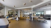 Located at 255 Stillwater Dr in Saskatoon, Saskatchewan, this dedicated exercise room offers a well-rounded fitness facility designed to support a variety of workout routines. The space features a durable rubber flooring surface throughout, providing excellent traction and cushioning suitable for both cardio and strength training activities.

The room is thoughtfully divided into distinct workout zones. The cardio area is equipped with a treadmill, elliptical trainer, stationary upright bike, and a recumbent cycle, positioned along a row of windows that allow natural light to fill the space. The strength training section includes a multi-station cable machine, a comprehensive dumbbell rack with a full range of free weights, and a variety of benches for targeted exercises.

Full-length mirrors line multiple walls, enhancing the sense of openness and allowing users to monitor their form during workouts. Recessed track lighting illuminates the space evenly, while structural columns define areas without impeding movement. A wall-mounted television adds convenience for those who enjoy watching content while exercising. Additional amenities visible in the space include a locker or storage area and posted informational signage. The clean, white walls and bright overall aesthetic contribute to an energizing and functional environment.