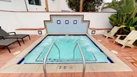 This outdoor amenity space showcases a well-appointed spa and lounge area designed in a Spanish Colonial Revival style. The centerpiece is a generously sized hot tub with bubbling turquoise water, framed by a striking decorative tile backsplash in blue and white with ornate patterned borders. Stainless steel entry rails provide safe access, and the surrounding deck is finished in warm terracotta brick pavers that complement the white stucco architecture throughout.

An arched colonnade runs along one side of the patio, fitted with classic black lantern-style wall sconces that provide ambient evening lighting. Colorful Adirondack chairs in teal and natural wood tones are arranged near the spa and beneath the archways, offering relaxed seating. Additional cushioned chaise lounges are positioned nearby for sunbathing. Lush tropical plantings, including tall palm trees, banana plants, and potted greenery, enhance the resort atmosphere.

The space connects visually to an adjacent pool area, visible beyond the colonnade arches with blue umbrellas and open sky. Spa rules and capacity signage are clearly posted, noting a capacity of 13. The overall ambiance is warm, inviting, and distinctly Mediterranean in character.