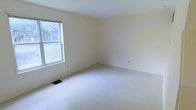 This two-story property presents a straightforward and functional layout spread across two floors, offering ample living space with a clean, neutral aesthetic throughout. The interiors are finished with light cream-colored walls that contribute to a bright, airy atmosphere, complemented by wall-to-wall carpeting that adds warmth and comfort underfoot. Natural light plays a significant role in the ambiance of the space, with windows framing views of surrounding greenery, bringing the outdoors in and enhancing the sense of openness in each room. The floor plan provides a versatile foundation, well-suited for a variety of furniture arrangements and personal styling preferences. Subtle architectural details include standard ceiling heights and straightforward room transitions that lend the space a cohesive, uncluttered feel. Floor vents integrated into the carpeted floors indicate a central HVAC system for year-round climate control. The overall condition of the property appears well-maintained, with smooth wall finishes and consistent flooring throughout. The neutral palette and abundant natural light make this a highly adaptable space, ready to accommodate a wide range of uses and interior design approaches across both levels.