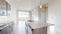 Located at 5909 Sharpsburg Dr in Madison, Wisconsin, this single-floor unit offers a clean, contemporary design with an open-concept layout that maximizes both functionality and natural light. The kitchen serves as the centerpiece of the space, anchored by a generous island topped with granite countertops and an undermount sink illuminated by a trio of pendant lights. Rich dark-toned cabinetry pairs with a classic white subway tile backsplash, while a full suite of stainless steel appliances — including a range, microwave, and refrigerator — completes the well-appointed cooking space. Light wood-tone flooring extends seamlessly throughout the unit, connecting the kitchen to the open living area. Floor-to-ceiling windows and a sliding glass door bathe the living space in natural light and provide direct access to a private balcony with outdoor views. The bedroom is bright and airy, benefiting from the same generous glazing and balcony access. The bathroom features granite countertops consistent with the kitchen, dark cabinetry with brushed nickel hardware, a large mirror, and a tub-shower combination, all finished with the same light flooring. The overall ambiance is modern, bright, and well-coordinated throughout.