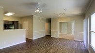 This single-floor space offers a clean, functional layout with a warm and inviting aesthetic. The main living area is finished with rich, dark hardwood-style flooring that contrasts beautifully against the neutral, earthy-toned walls. Crown molding adds a refined architectural touch, running along the upper edges of the walls to lend a polished look throughout. A ceiling fan with integrated lighting anchors the main living space, providing both comfort and illumination, while a track lighting fixture in the adjacent area offers additional directional light. White panel doors and crisp white baseboards create a bright, cohesive contrast against the warm wall tones. The space transitions seamlessly into additional rooms, which appear to feature carpeted flooring — a practical contrast to the hard flooring in the common areas. A half-wall divider subtly defines separate zones within the open layout without compromising the sense of openness. The overall ambiance is bright, airy, and move-in ready, with a straightforward design that lends itself well to a variety of furnishing styles. The combination of practical finishes, thoughtful architectural details, and a neutral palette makes this a versatile and comfortable space.
