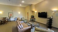 This single-floor space serves as a communal lounge or gathering area, designed with functionality and comfort in mind. The room features a dropped acoustic tile ceiling with recessed lighting, complemented by warm wall-mounted sconces that cast a welcoming ambient glow against the beige-toned walls. The space is anchored by a large leather sofa and an upholstered armchair arranged around a glass-top coffee table with a metal frame, creating a defined seating area suited for relaxed conversation or group gatherings. A large flat-screen television is mounted prominently on one wall, while an older CRT television on a corner stand provides an additional viewing option. The flooring is covered in neutral carpet that extends throughout the room, adding to the understated, practical aesthetic. A structural column with a tiered architectural detail adds a subtle design element near the center of the space. A doorway visible in the background suggests connectivity to adjacent rooms or corridors. Overall, the space presents a casual, utilitarian atmosphere well-suited for a shared or communal environment.