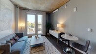 This contemporary single-floor residence showcases a sophisticated industrial-modern aesthetic defined by signature exposed concrete ceilings that span throughout the space, paired with warm hardwood flooring for a balanced, stylish contrast. The open layout flows naturally between the key living areas, creating a cohesive and functional environment.

The living room is bright and inviting, featuring large French doors that open to a private balcony with expansive city and treetop views. A wall-mounted split-system unit provides climate control, while the concrete ceiling overhead reinforces the urban character of the space.

The kitchen is efficiently designed with warm wood cabinetry, light countertops, and a full suite of stainless steel appliances including a gas range, built-in microwave, refrigerator, and dishwasher. Track lighting illuminates the workspace, and the layout maximizes usability within a streamlined footprint.

The bedroom is a serene retreat, anchored by a large picture window framing striking city skyline and greenery views. The exposed concrete ceiling continues here, adding architectural texture, while soft ambient lighting creates a calm and restful atmosphere.

The bathroom is cleanly finished with large-format gray tile surrounding a full soaking tub and shower combo, complemented by chrome fixtures and wood-tone flooring for a refined, spa-like feel.
