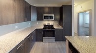 This single-floor residence offers a thoughtfully designed layout with modern finishes throughout. The kitchen is a standout feature, showcasing rich dark wood cabinetry extending to the ceiling, granite countertops, a subway tile backsplash, and stainless steel appliances including a range, microwave, and refrigerator. A kitchen island with pendant lighting and an undermount sink bridges the kitchen to the open living area, which features light hardwood-style flooring, a ceiling fan, and a glass door leading to a private balcony that brings in ample natural light. The carpeted bedrooms are generously sized, each equipped with a ceiling fan, a window with blinds, and access to a walk-in closet. The bathrooms are finished in a clean, bright white palette — one featuring a soaking tub with a curved shower rod and built-in shelving, while the second includes a tub-shower combo, tile flooring, and a convenient stacked washer and dryer unit. The overall ambiance of the space is clean, contemporary, and functional, with neutral tones and consistent finishes that create a cohesive and welcoming atmosphere throughout.