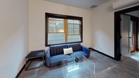 This single-floor unit showcases a cohesive modern design anchored by polished concrete floors that flow seamlessly throughout the entire space, lending an industrial yet refined character to every room.

The kitchen is a well-appointed culinary hub, outfitted with stainless steel appliances including a gas range, refrigerator, dishwasher, and built-in microwave. Two-toned cabinetry — dark lower cabinets paired with taupe upper cabinets — frames a clean white subway tile backsplash and light-toned countertops. A compact center island with bar seating provides additional prep space and casual dining.

The living area benefits from a large, wood-framed picture window with dark trim, allowing natural light to filter into the room. Contemporary furnishings, including a channel-tufted blue sofa and a clear acrylic coffee table, complement the understated, neutral walls.

The bathroom features a dark-vanity with an integrated sink, a framed mirror, and a shower enclosed by a curtain. Tall grey storage cabinetry offers ample organizational space. The continued use of polished concrete flooring unifies the bathroom with the rest of the unit, reinforcing the space's consistent industrial-modern aesthetic.