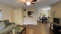 This two-floor residence offers a thoughtfully designed layout that maximizes both functionality and comfort. The main living area is open-concept, seamlessly connecting the living room, dining area, and kitchen into one cohesive space. Rich hardwood-style flooring runs throughout, adding warmth and continuity to the interior.

The living room is anchored by a ceiling fan with integrated lighting, providing both comfort and illumination. A set of built-in open shelving units lines one wall, offering ample display and storage space alongside a louvered-door closet. Neutral, warm-toned walls throughout create a welcoming and versatile backdrop.

The kitchen features dark cabinetry with a stainless steel microwave and appliances, accessible via a convenient pass-through opening that connects it to the main living space. This design enhances the sense of openness while keeping the kitchen functional and accessible.

The dining area is positioned adjacent to the kitchen and living room, accommodating a full dining set with ease. Decorative framed artwork adorns the walls, contributing to the polished aesthetic of the space.

With two floors, the residence provides additional living or sleeping areas beyond the main level, offering versatility and separation between living and private spaces. The overall ambiance is clean, bright, and practical.