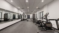 Located at 22 Schuyler Pl in Morristown, New Jersey, this fitness center offers a generously sized, single-floor exercise room designed to support a comprehensive workout experience. The space features a clean, modern aesthetic with crisp white walls, a suspended drop-tile ceiling with recessed lighting, and durable rubber flooring throughout — a practical and professional finish suited for high-traffic use.

The strength training area is anchored by a multi-functional cable machine and a full smith/power rack system paired with an adjustable bench, all positioned alongside a large wall-mounted mirror that enhances the sense of space and allows for proper form checks during workouts. A dedicated dumbbell rack offers a range of free weights for varied training.

The cardio zone lines the windowed wall, housing multiple treadmills, an elliptical, a stationary bike, and a rowing machine, providing ample options for cardiovascular training. Multi-pane black-framed windows allow natural light to filter into the room, creating a bright and energizing atmosphere.

Access is provided through both a set of double doors and a glass-paneled entry door, contributing to an open and welcoming flow. The overall layout is thoughtfully organized, offering distinct zones for strength and cardio training within a spacious, well-lit environment.