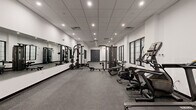 Located at 22 Schuyler Pl in Morristown, New Jersey, this fitness center offers a generously sized, single-floor exercise room designed to support a comprehensive workout experience. The space features a clean, modern aesthetic with crisp white walls, a suspended drop-tile ceiling with recessed lighting, and durable rubber flooring throughout — a practical and professional finish suited for high-traffic use.

The strength training area is anchored by a multi-functional cable machine and a full smith/power rack system paired with an adjustable bench, all positioned alongside a large wall-mounted mirror that enhances the sense of space and allows for proper form checks during workouts. A dedicated dumbbell rack offers a range of free weights for varied training.

The cardio zone lines the windowed wall, housing multiple treadmills, an elliptical, a stationary bike, and a rowing machine, providing ample options for cardiovascular training. Multi-pane black-framed windows allow natural light to filter into the room, creating a bright and energizing atmosphere.

Access is provided through both a set of double doors and a glass-paneled entry door, contributing to an open and welcoming flow. The overall layout is thoughtfully organized, offering distinct zones for strength and cardio training within a spacious, well-lit environment.