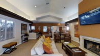 This single-floor clubhouse is thoughtfully designed to serve as a comfortable and inviting community gathering space. The centerpiece of the interior is a spacious open lounge area highlighted by a striking vaulted ceiling that adds volume and architectural character to the room. The soaring roofline, accented by clean white surfaces and recessed lighting, creates a bright and airy ambiance throughout.

The lounge is furnished with a large, plush cream sofa adorned with vibrant accent pillows in warm tones of gold, orange, and rust, complemented by rich leather armchairs and a sleek coffee table. A warm terracotta-toned feature wall provides a grounding contrast to the lighter surrounding walls, and curated wall art adds a refined, modern touch to the décor.

Adjacent to the main seating area, a kitchenette and bar counter with barstool seating offers a convenient space for casual dining or social gatherings. The area is framed by windows that allow natural light to filter in, enhancing the welcoming atmosphere. Dark wood trim details along the upper walls add a sophisticated finishing element that ties the overall design together, creating a space that is both functional and aesthetically inviting for residents and guests alike.