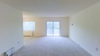 This single-floor space features an open and airy living area finished with wall-to-wall carpeting in a neutral tone, contributing to a clean and understated aesthetic throughout. The room is bathed in natural light, sourced from two windows — a sizable horizontal window fitted with blinds on one side, and a central sliding glass door that opens directly onto a private balcony. The balcony, enclosed by a railing, overlooks mature trees, offering a pleasant outdoor view and a connection to the surrounding greenery. The walls are painted in a soft, warm white, enhancing the brightness of the interior and creating a versatile backdrop for a variety of décor styles. A baseboard heating unit runs along the base of the exterior wall, providing functional climate control. The open layout of the living area allows for flexible furniture arrangement, making the space adaptable to different uses. The overall ambiance is calm and well-lit, with the abundance of natural light and neutral finishes giving the space a welcoming and move-in-ready character.