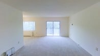 This single-floor space features an open and airy living area finished with wall-to-wall carpeting in a neutral tone, contributing to a clean and understated aesthetic throughout. The room is bathed in natural light, sourced from two windows — a sizable horizontal window fitted with blinds on one side, and a central sliding glass door that opens directly onto a private balcony. The balcony, enclosed by a railing, overlooks mature trees, offering a pleasant outdoor view and a connection to the surrounding greenery. The walls are painted in a soft, warm white, enhancing the brightness of the interior and creating a versatile backdrop for a variety of décor styles. A baseboard heating unit runs along the base of the exterior wall, providing functional climate control. The open layout of the living area allows for flexible furniture arrangement, making the space adaptable to different uses. The overall ambiance is calm and well-lit, with the abundance of natural light and neutral finishes giving the space a welcoming and move-in-ready character.