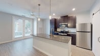 This well-appointed single-floor residence showcases a thoughtfully designed open-concept layout that seamlessly connects the kitchen and living areas. The kitchen is a standout feature, anchored by a large center island with a gray quartz-style countertop, an undermount sink with a brushed gold faucet, and stylish pendant lighting overhead. Rich dark cabinetry provides generous storage and pairs elegantly with stainless steel appliances, including a range, microwave, dishwasher, and refrigerator. Light wood-style flooring runs throughout the space, creating a warm, cohesive aesthetic. A ceiling fan enhances the comfort of the living area, which also benefits from natural light through a glass door leading to the outdoors. A distinctive sliding barn door adds a modern design touch and leads to an additional room or storage area with built-in shelving. The bedroom is spacious and bright, featuring a built-in closet and direct sightlines to the kitchen. The bathroom is clean and functional, offering a tiled tub-shower combination with brushed gold fixtures. Overall, the space presents a fresh, contemporary feel with consistent finishes and a practical, flowing floor plan.