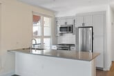 Located at 1650 S Delaware St, Unit 206 in San Mateo, CA, this thoughtfully designed single-floor residence offers a clean, contemporary aesthetic throughout. The open-concept kitchen is a standout feature, showcasing white shaker-style cabinetry with glass-front upper panels, a classic subway tile backsplash, stainless steel appliances including a range, microwave, dishwasher, and refrigerator, and a central island with a contrasting countertop — all set atop warm wood-tone flooring. The living area extends naturally from the kitchen and is bathed in natural light through large windows and a glass door that opens to an outdoor space with views of surrounding greenery. The bedrooms are carpeted for comfort and feature ceiling fans, natural light from well-placed windows, and a clean, neutral palette. The primary bathroom is generously proportioned, offering a walk-in shower with glass enclosure, a floating vanity with white quartz countertop and black hardware, and access to a walk-in closet with built-in shelving. A secondary full bathroom mirrors this refined aesthetic with a shower-tub combination and matching vanity finishes. Throughout the residence, crisp white walls, recessed lighting, and consistent wood-tone flooring create a cohesive and modern living environment.